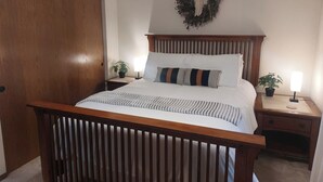 A neatly arranged bedroom with a wooden bed frame, a headboard with a wreath, two bedside tables with lamps, and a potted plant on each side.