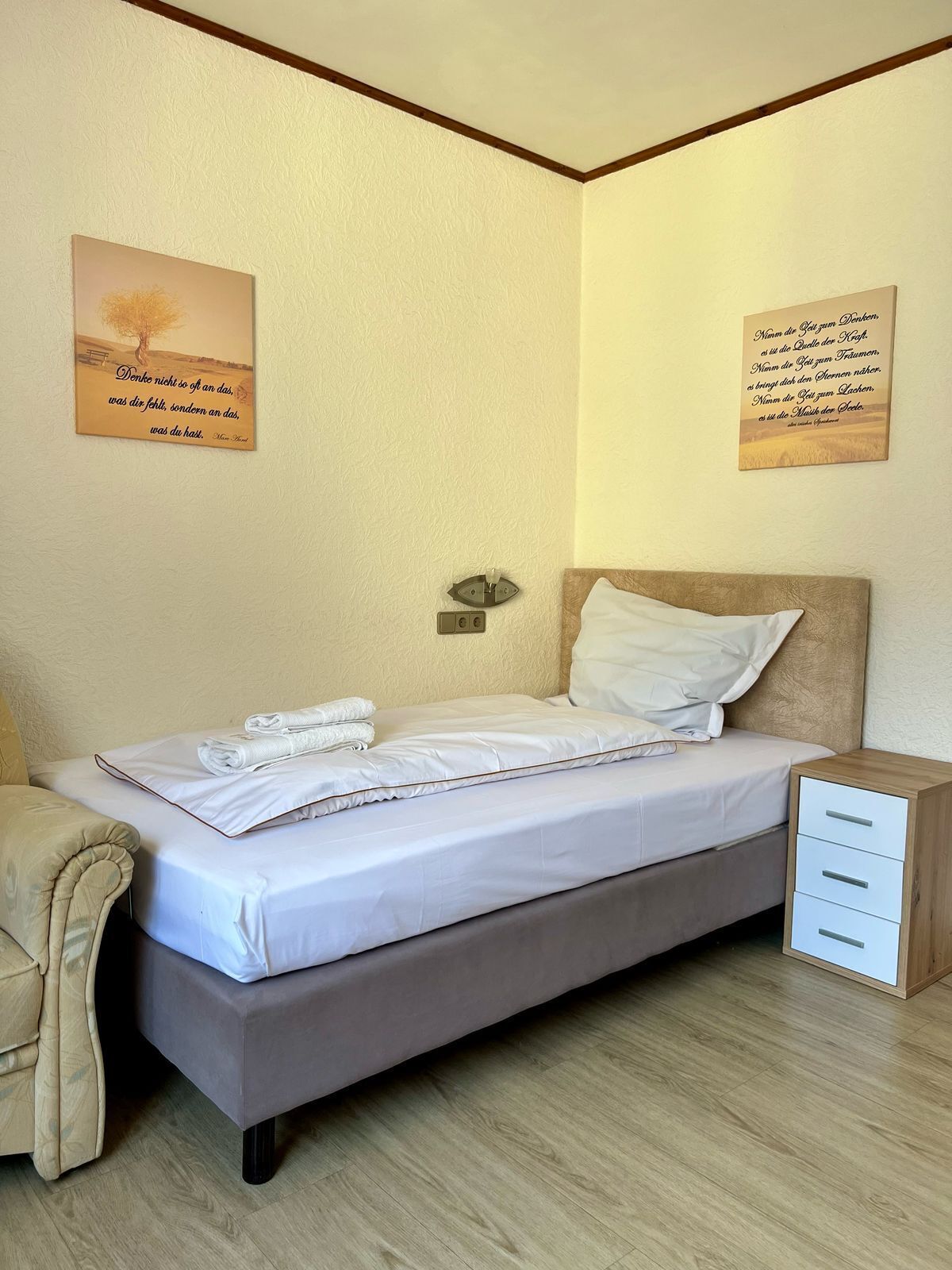 A neatly made bed with white linens and a folded towel, a bedside table with a lamp, and two wall-mounted artworks.