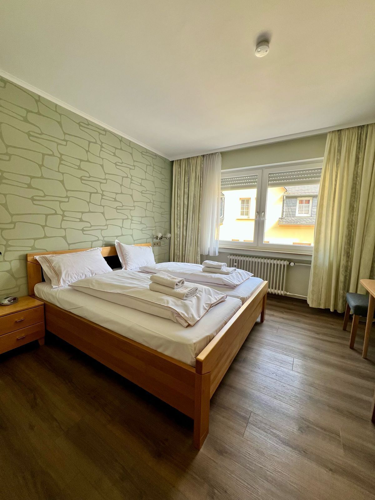 Un lit double bien fait, avec du linge de lit blanc, une table de chevet en bois, une petite table avec une chaise et une fenêtre avec des rideaux.