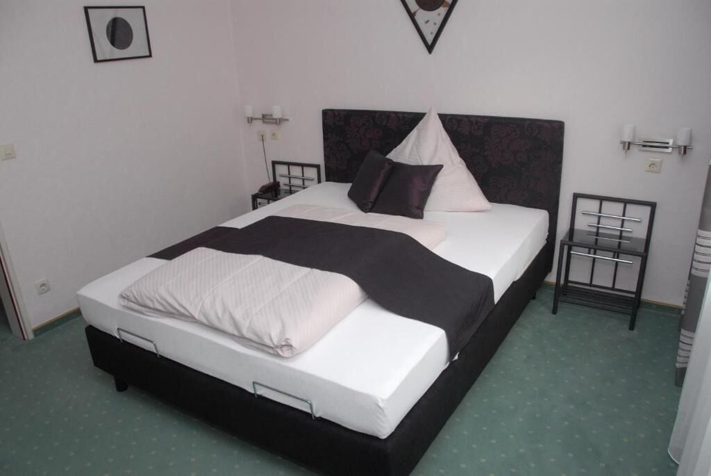 A neatly made bed with a black headboard, white bedding, and a black throw blanket. There are two bedside tables with lamps, and a small picture frame on the wall.