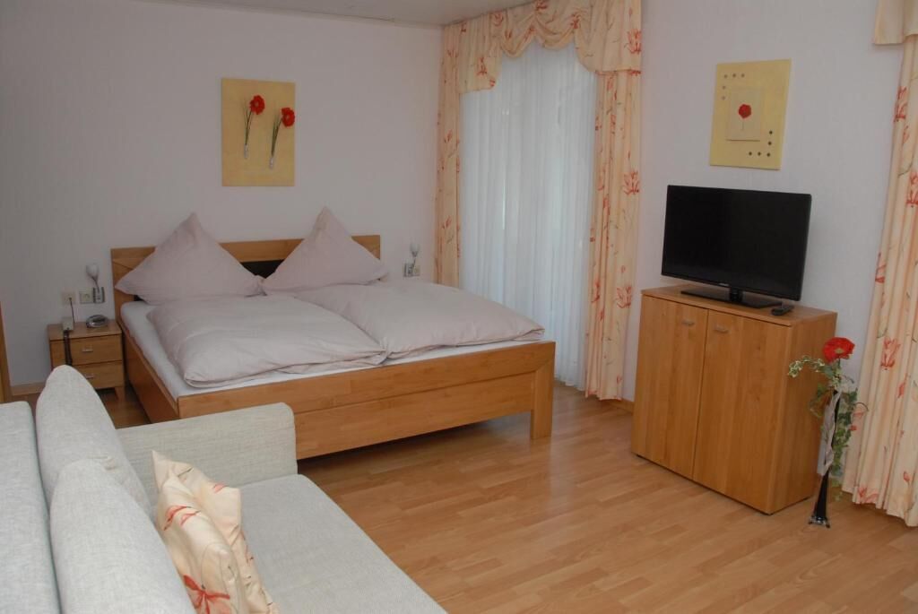 Ein Schlafzimmer mit einem hölzernen Bett, einem Fernseher auf einer Holzkommode, einem Nachttisch und einem blumigen Vorhang.