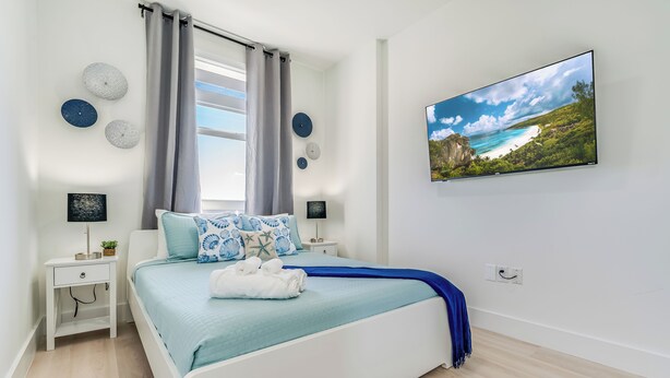 Une chambre à coucher avec un lit, une table de chevet, un téléviseur fixé au mur et une fenêtre avec des rideaux.