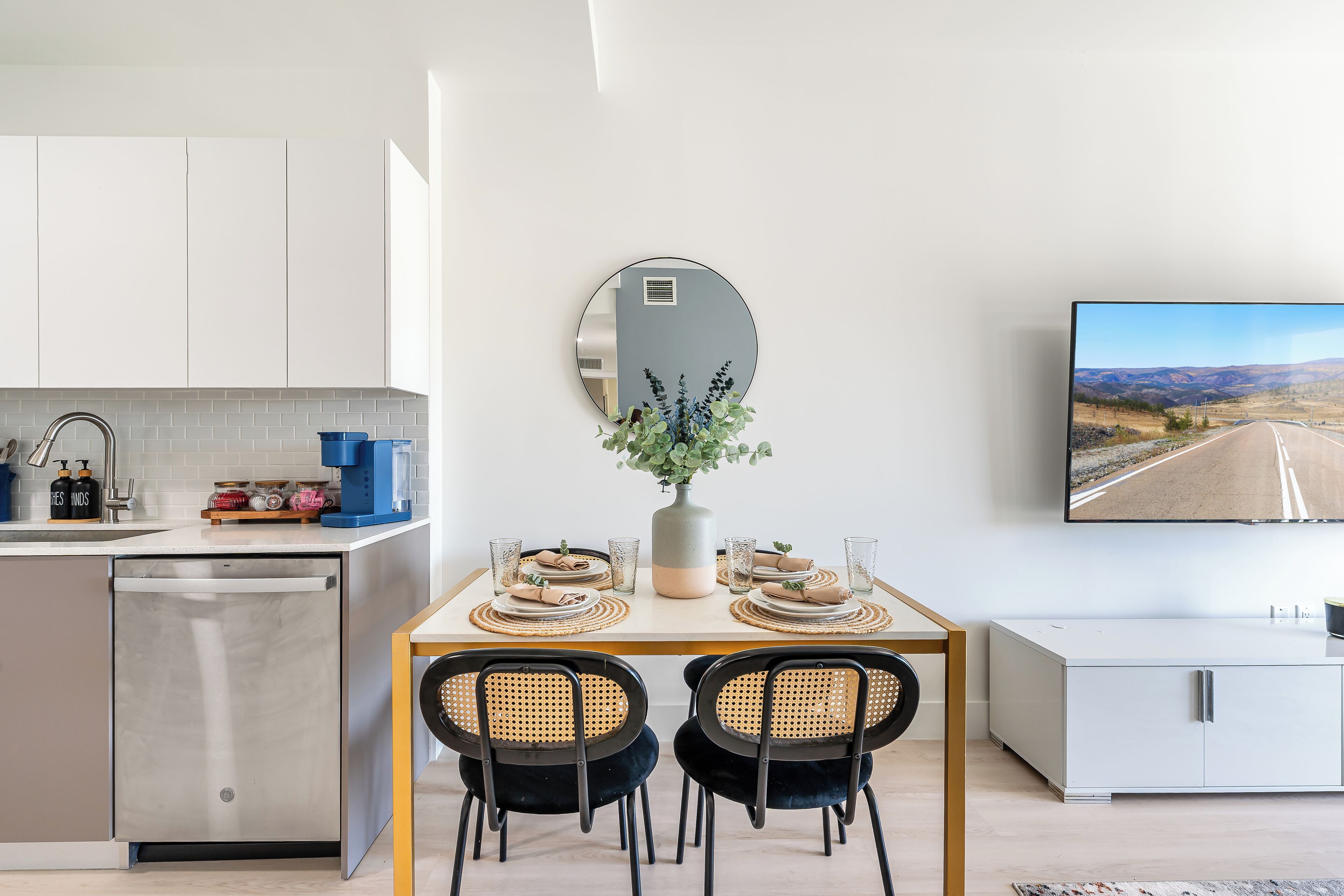 Une cuisine moderne équipée d’un lave-vaisselle, d’une table à manger avec deux chaises, d’un vase agrémenté de plantes et d’un téléviseur à écran plat fixé au mur.
