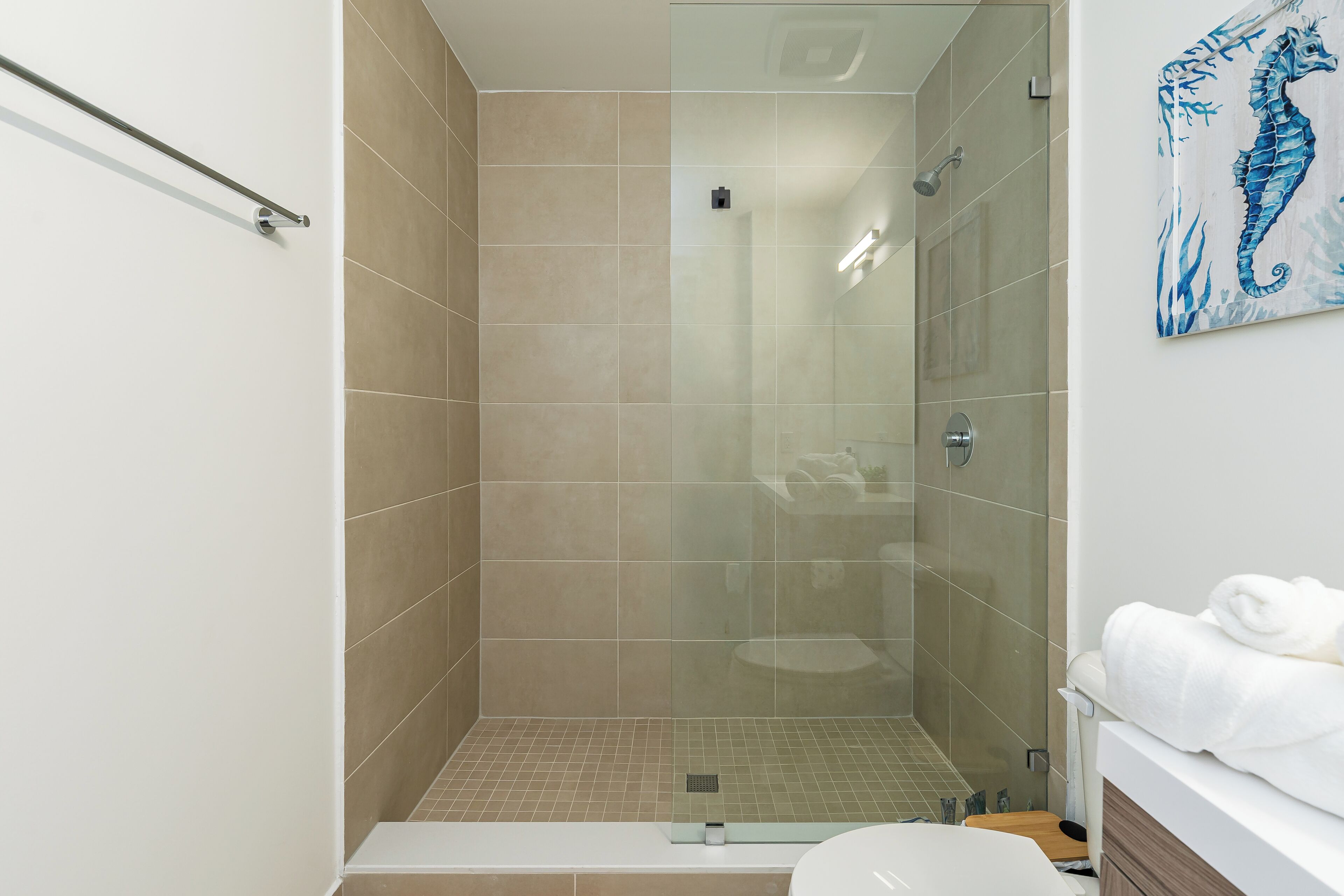 A modern bathroom with a glass-enclosed shower, beige tiles, a white sink, and a seahorse artwork on the wall.