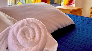 A bed with a blue quilt and white pillows, a rolled white towel, and yellow curtains. - Hotel Minnesota (San Lucas Tolimán)