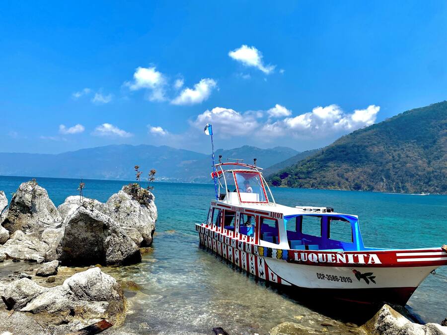 A boat named "LUQUENITA" docked at a rocky shore with clear water and mountains in the background.