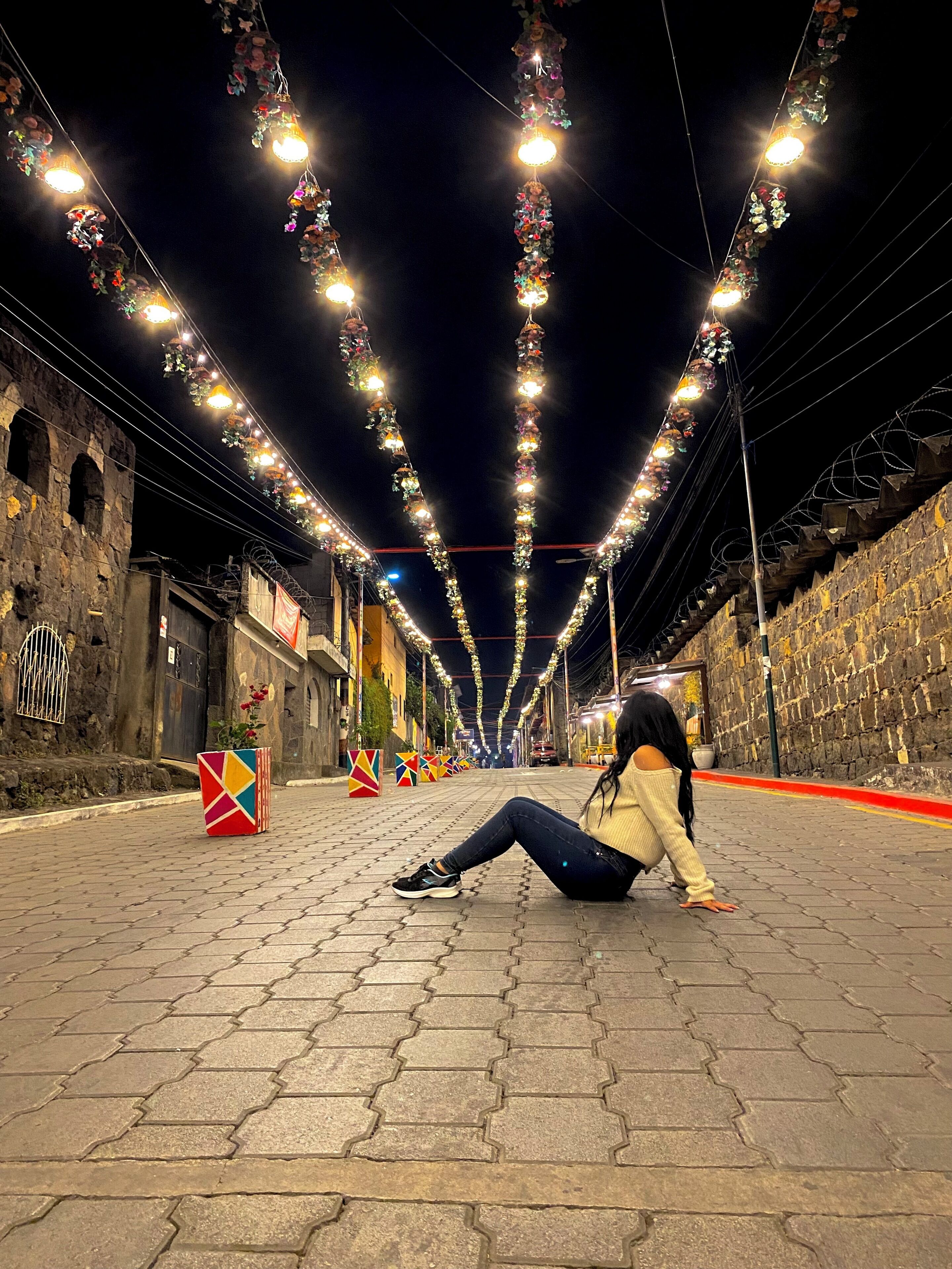 A street at night with decorative lights and a person sitting on the ground.
