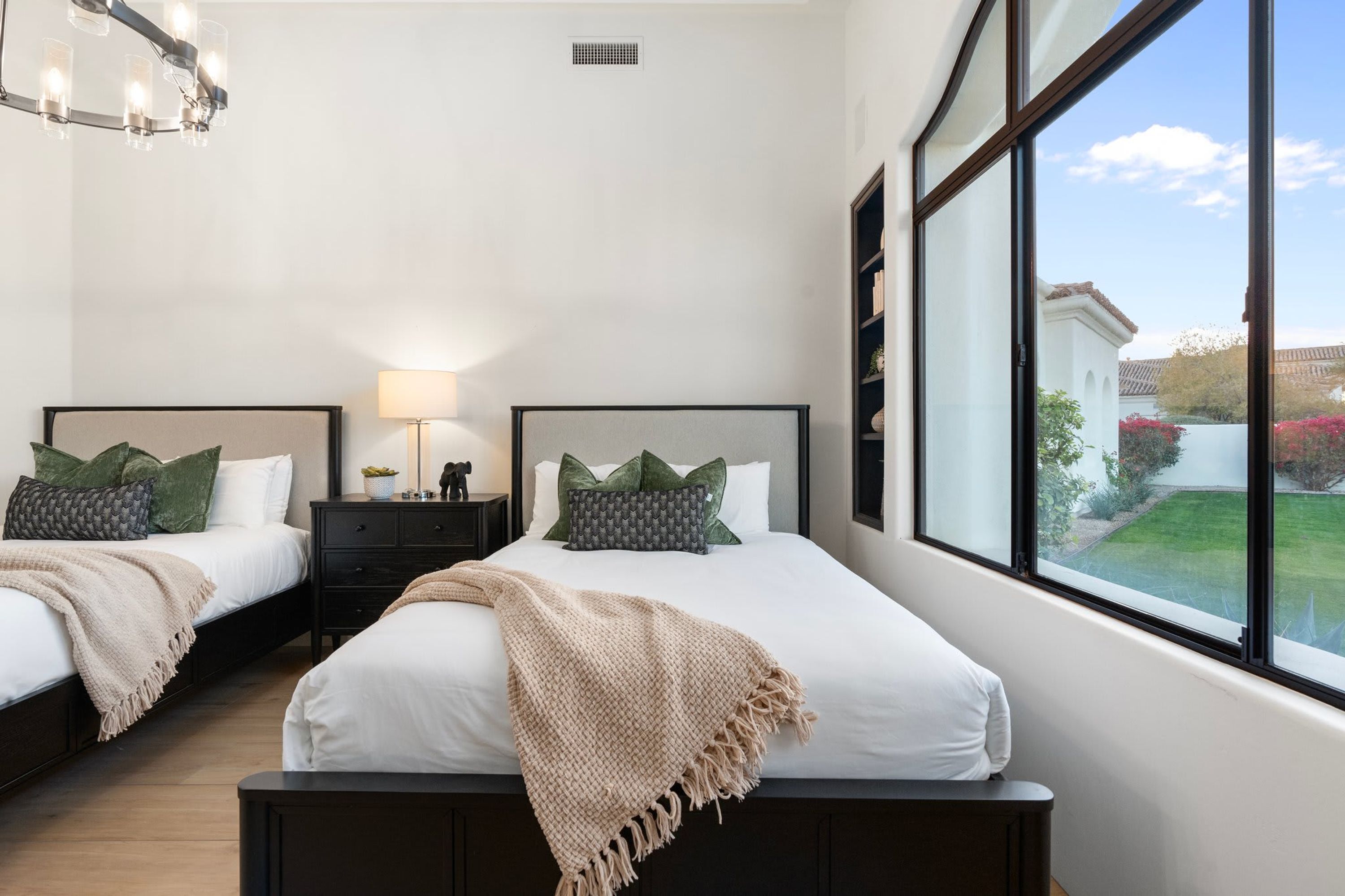 A bedroom with two beds, a nightstand, a lamp, and a large window with a view of a garden.