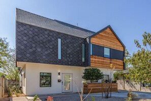 Una casa moderna de dos pisos con techo de tejas oscuras, sección frontal de madera y un pequeño jardín con plantas y un árbol.