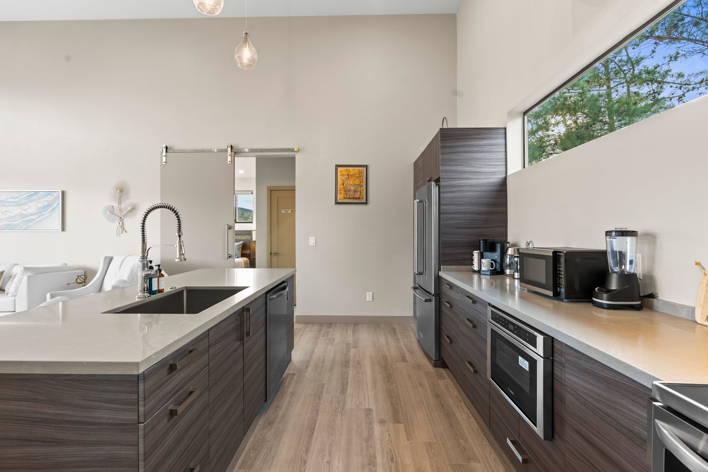 A modern kitchen with a large island, stainless steel appliances, and a view of trees through a large window.