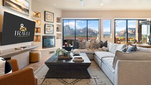 A modern living room with a large flat-screen TV, comfortable seating, a coffee table with decorative items, and a view of mountains through large windows.