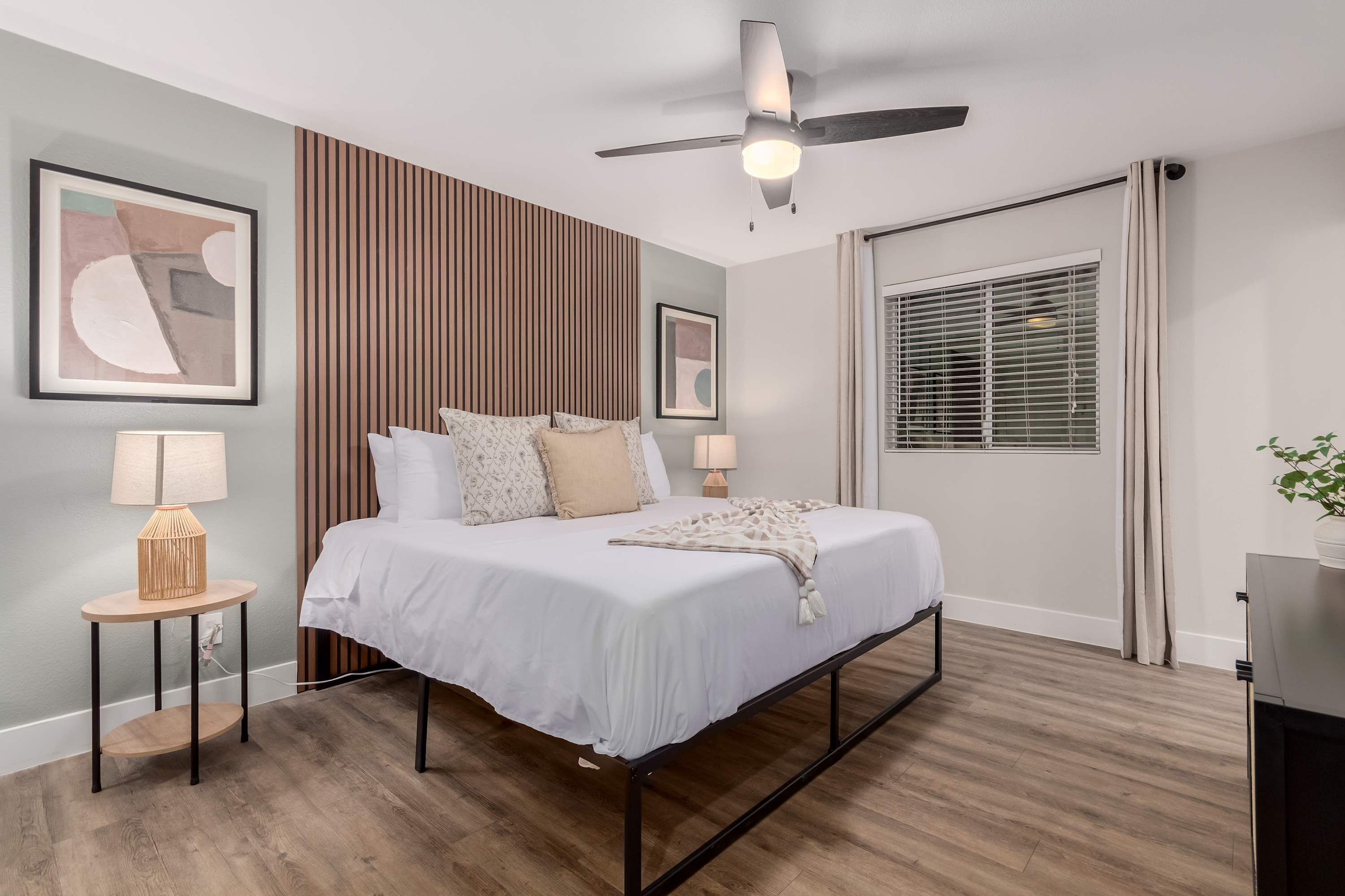A modern bedroom with a bed, bedside lamps, a ceiling fan, and a window with blinds.