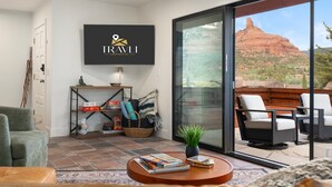A modern living room with a flat-screen TV displaying the TRAVLI logo, a comfortable seating area with a sofa and chairs, a coffee table with books, and a large sliding glass door leading to an outdoor patio with a view of a red rock formation. - NEW Redrock Experience Views Firepit Hottub (Sedona)
