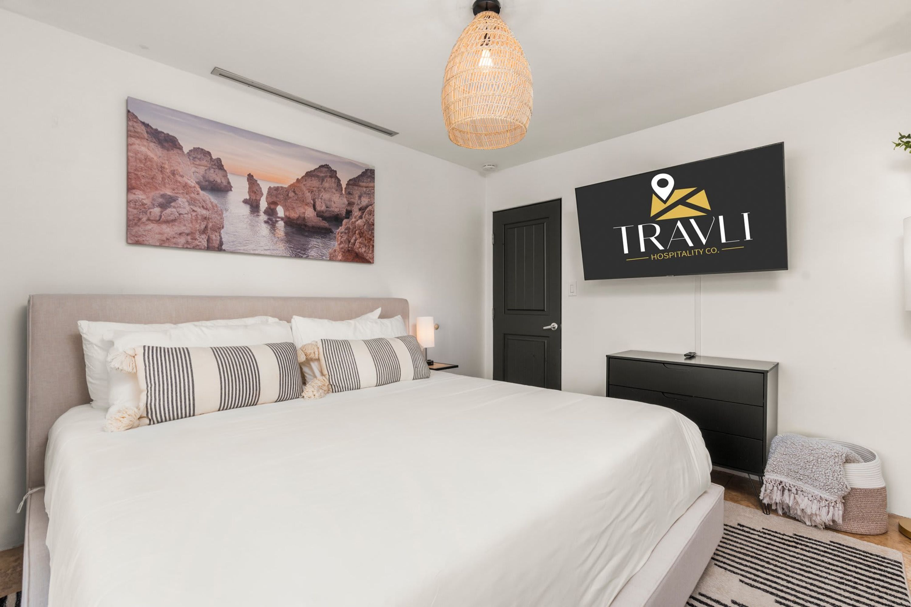 A bedroom with a bed, a TV mounted on the wall, a door, and a painting of a coastal landscape.