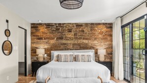 Une chambre avec un mur en pierre, un lit avec une literie blanche, deux tables de chevet avec des lampes, un banc et une grande fenêtre avec des rideaux.
