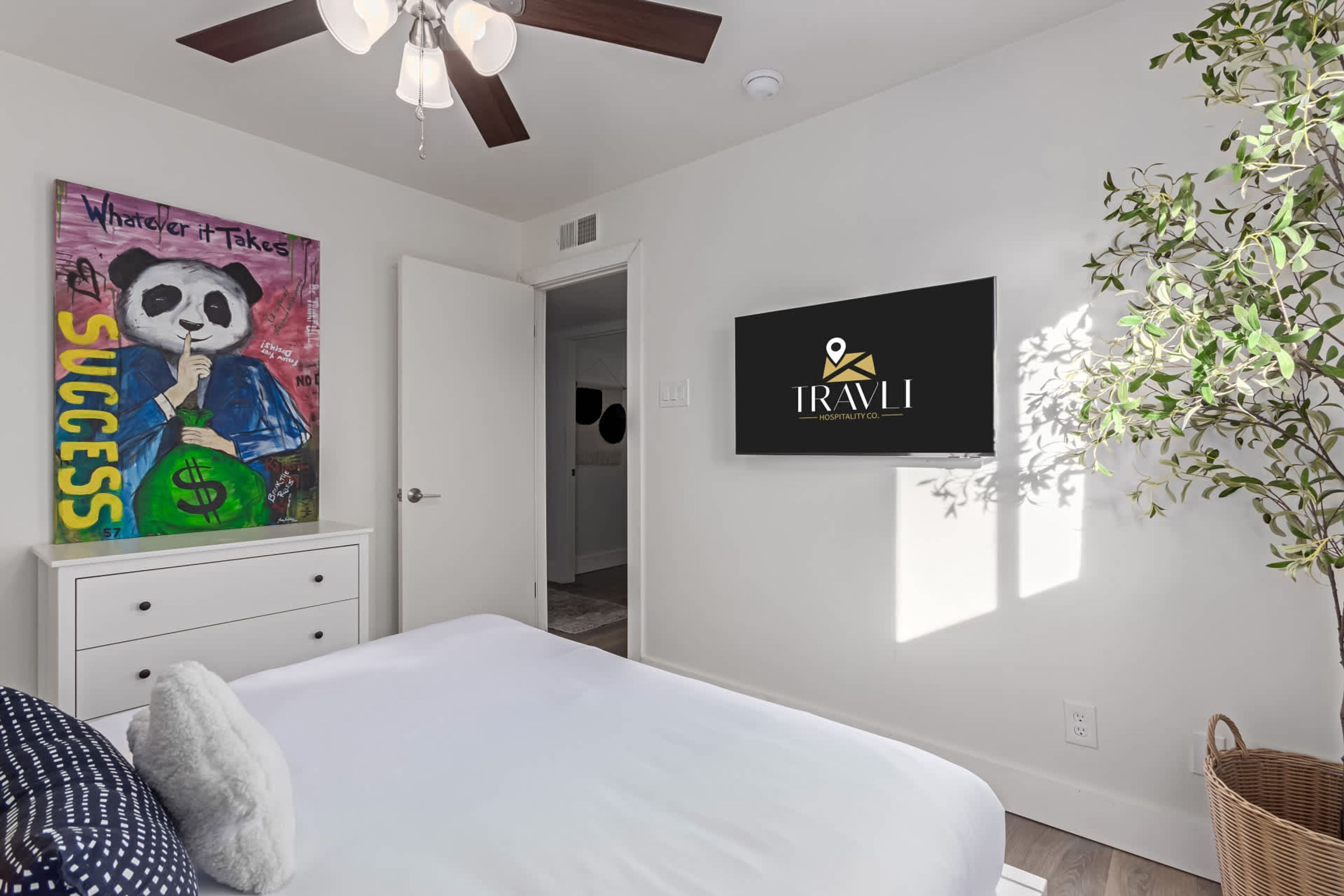 A bedroom with a bed, a ceiling fan, a wall-mounted TV, a poster, a door, and a potted plant.