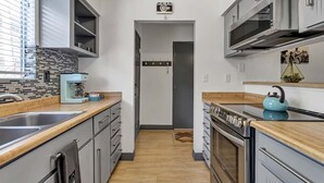 Una cocina pequeña con microondas, estufa y lavamanos. Hay una ventana con persianas y una puerta que conduce a otra habitación.