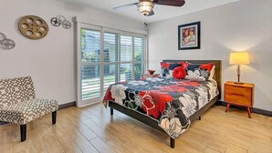 Un dormitorio con cama, ventilador de techo, ventana con persianas, una silla y una mesita de noche con lámpara.