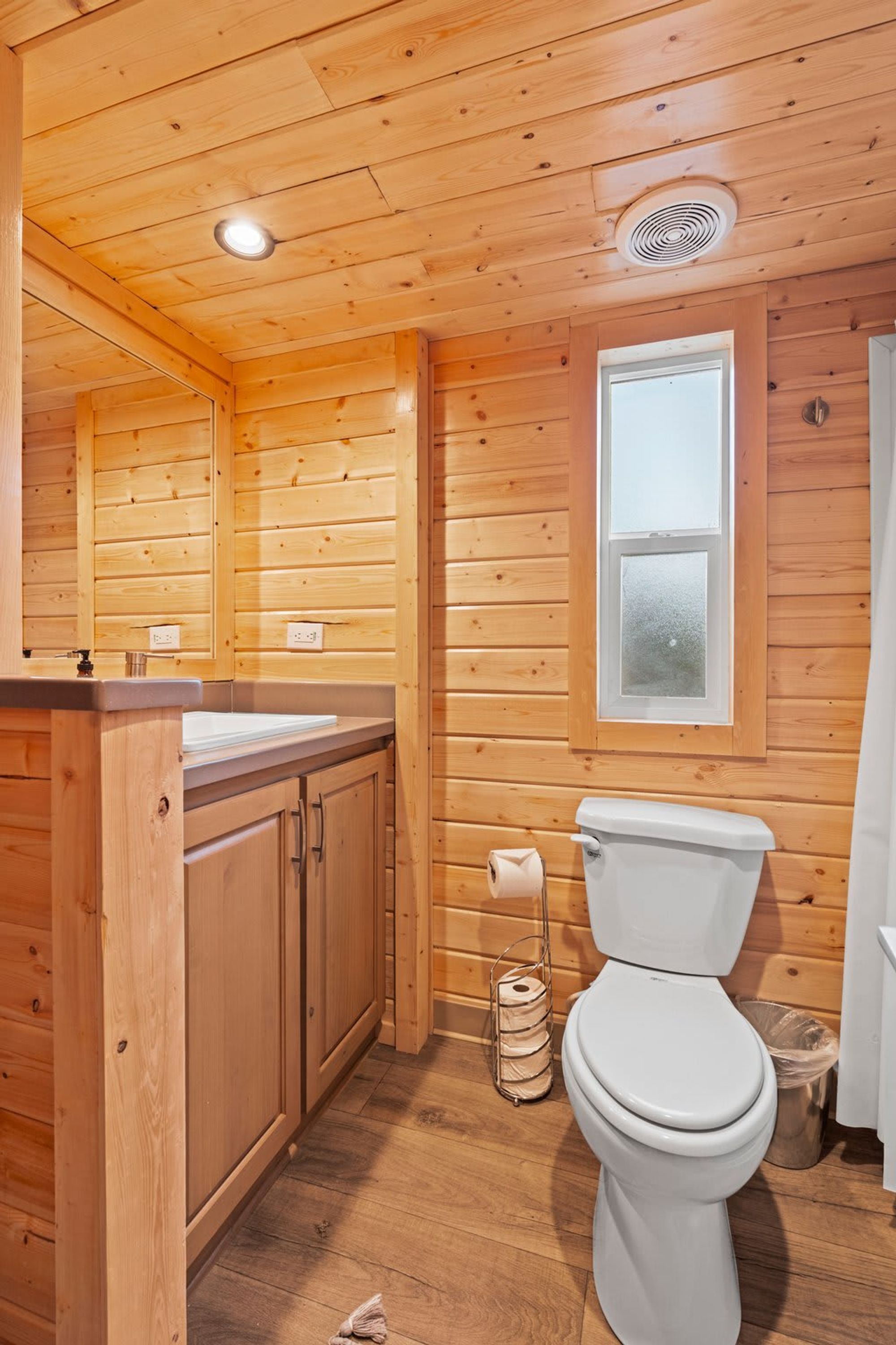 A wooden cabin bathroom with a toilet, sink, and window.