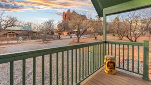 A wooden deck with a green railing, a rustic wooden sculpture, and a view of a rocky mountain and buildings in the distance. - NEW Moondance Majestic Mini Cabin Cathedral Rock (Sedona)