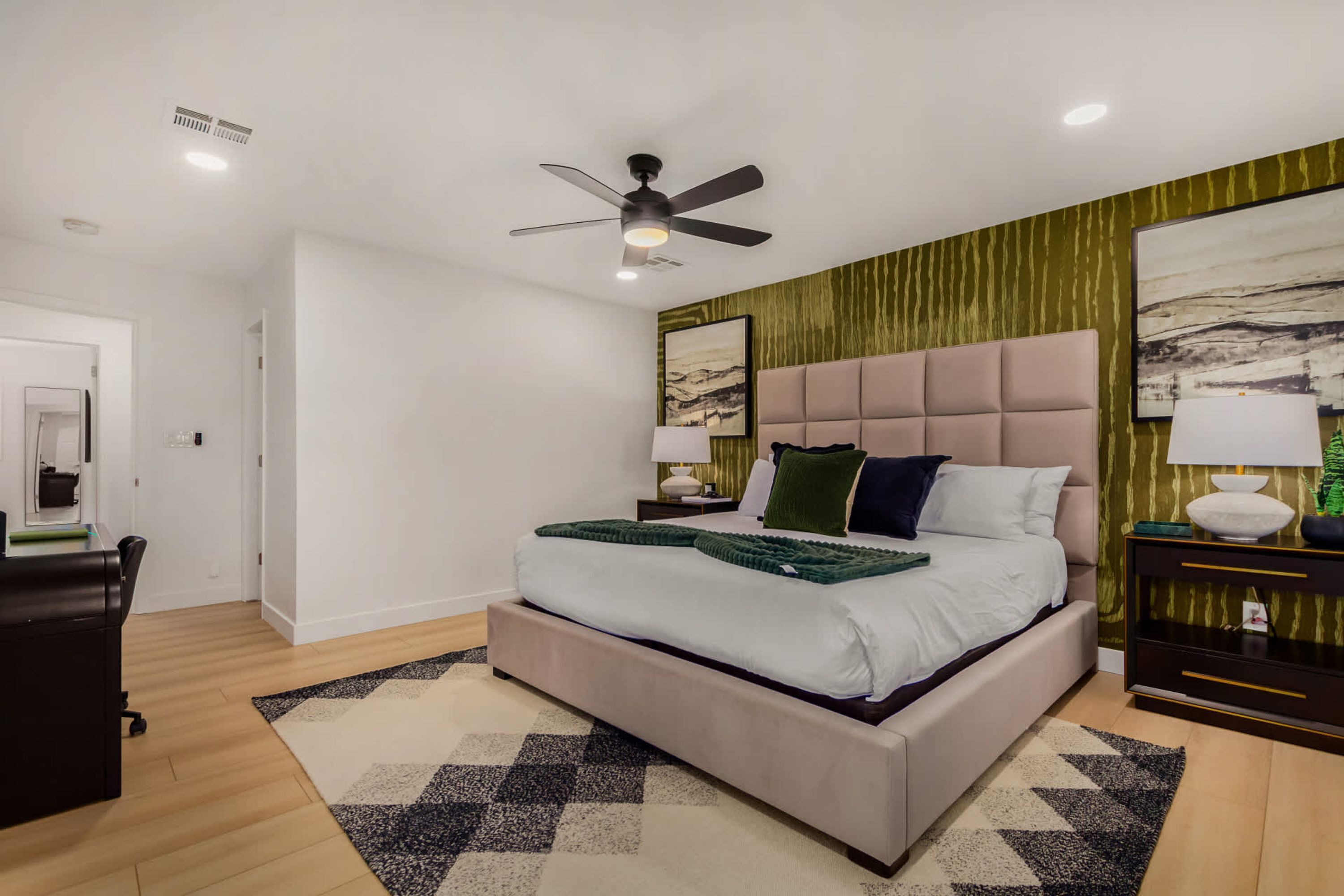 A modern bedroom with a large bed, a ceiling fan, a nightstand with a lamp, and a wall with abstract art.