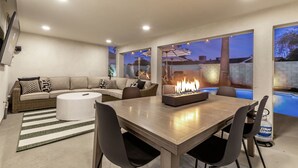 A modern living room with a wooden dining table, comfortable seating, a fireplace, and a view of a swimming pool at dusk.