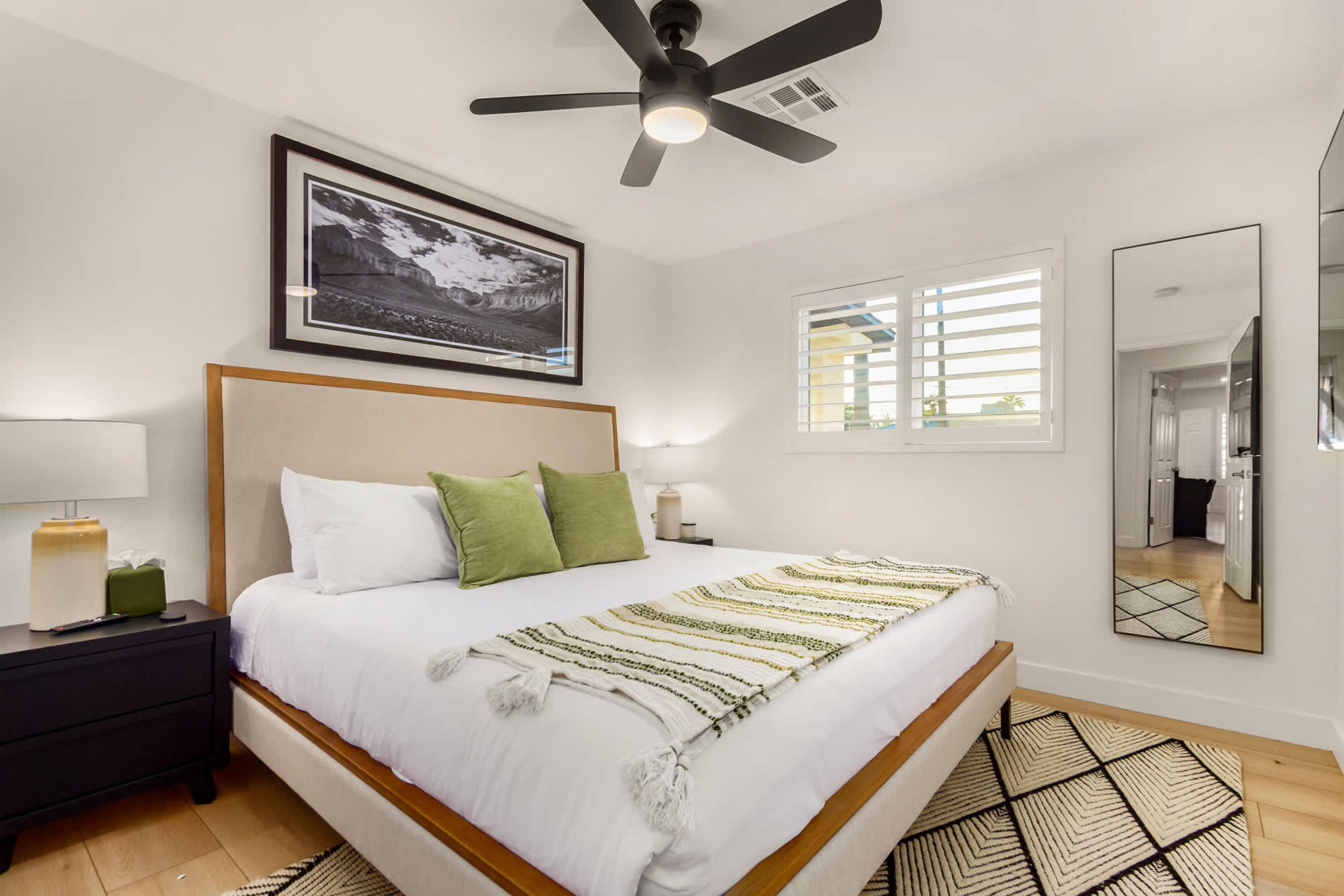 A neatly arranged bedroom with a bed, bedside tables, a ceiling fan, a framed picture, a window with blinds, and a mirror.