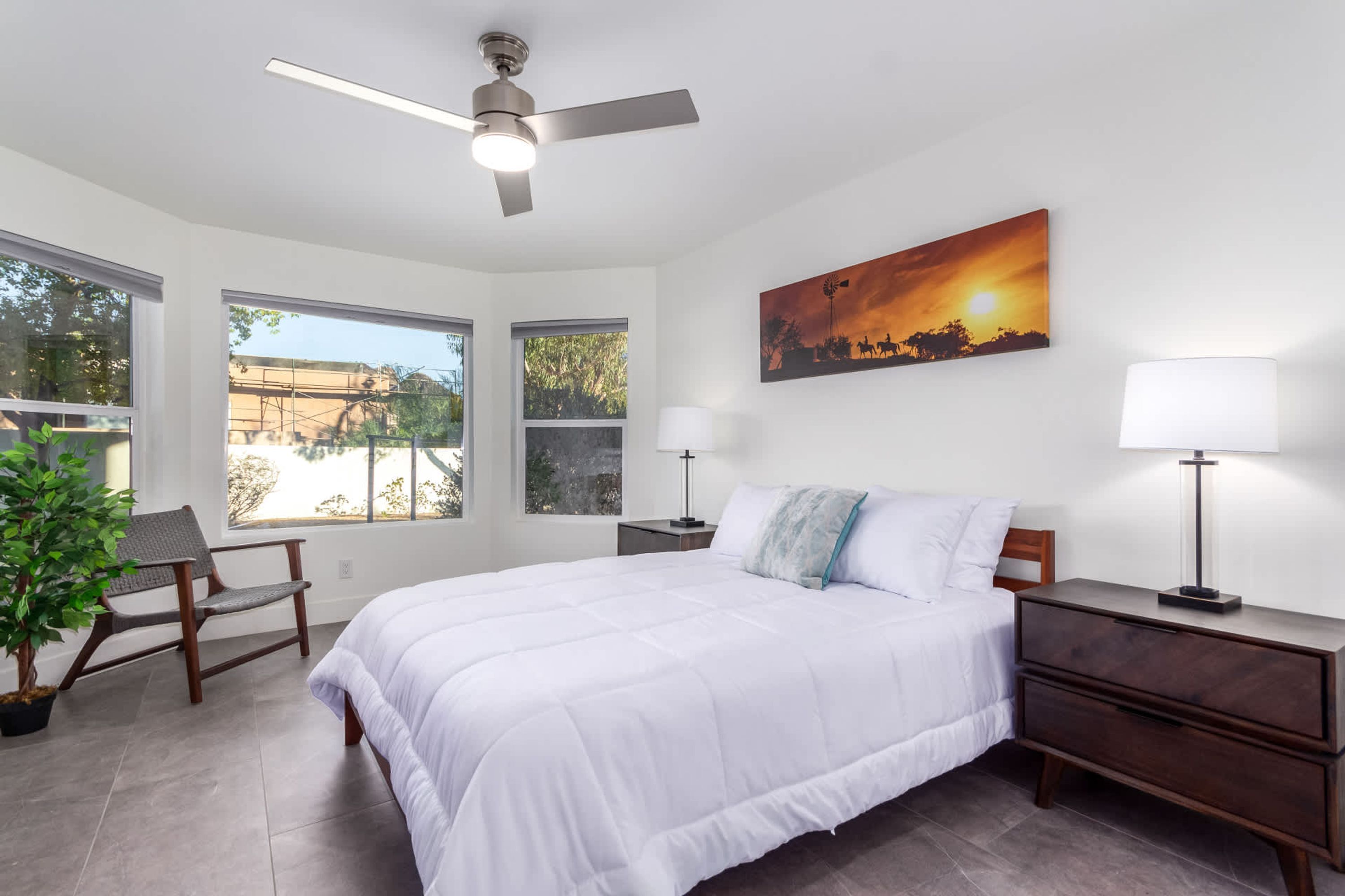 Quarto com cama, duas mesas de cabeceira, ventilador de teto, janela, cadeira e planta em vaso.