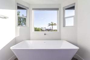 A modern bathroom with a freestanding bathtub, large windows with a view of palm trees and a clear sky.