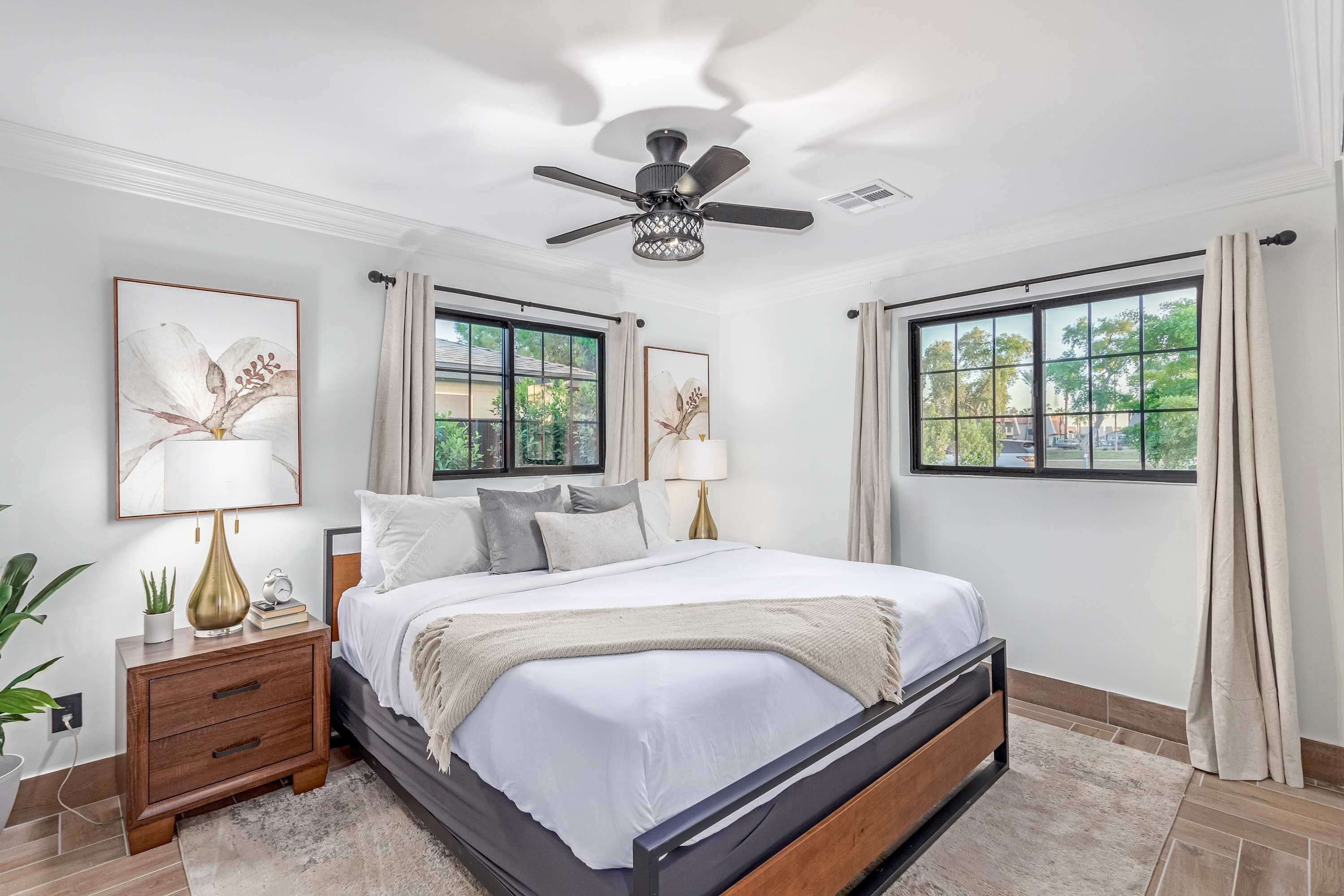 A bedroom with a bed, bedside tables, a ceiling fan, and two windows with curtains.