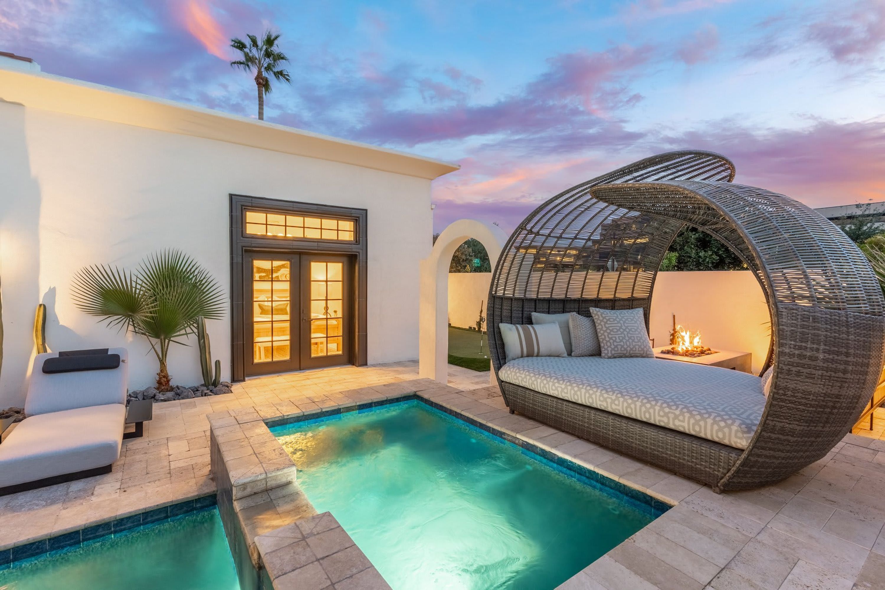 A modern outdoor pool area with a lounge chair, a wicker sofa, and a well-lit entrance.