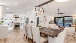 A modern kitchen with white cabinets, a wooden dining table, and a chandelier. - NEW The Kensington Custom Pool Al-fresco Living (Scottsdale)