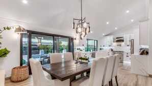 A modern kitchen with white cabinets, a stainless steel stove, and a dining area with a wooden table and white chairs. - NEW The Kensington Custom Pool Al-fresco Living (Scottsdale)
