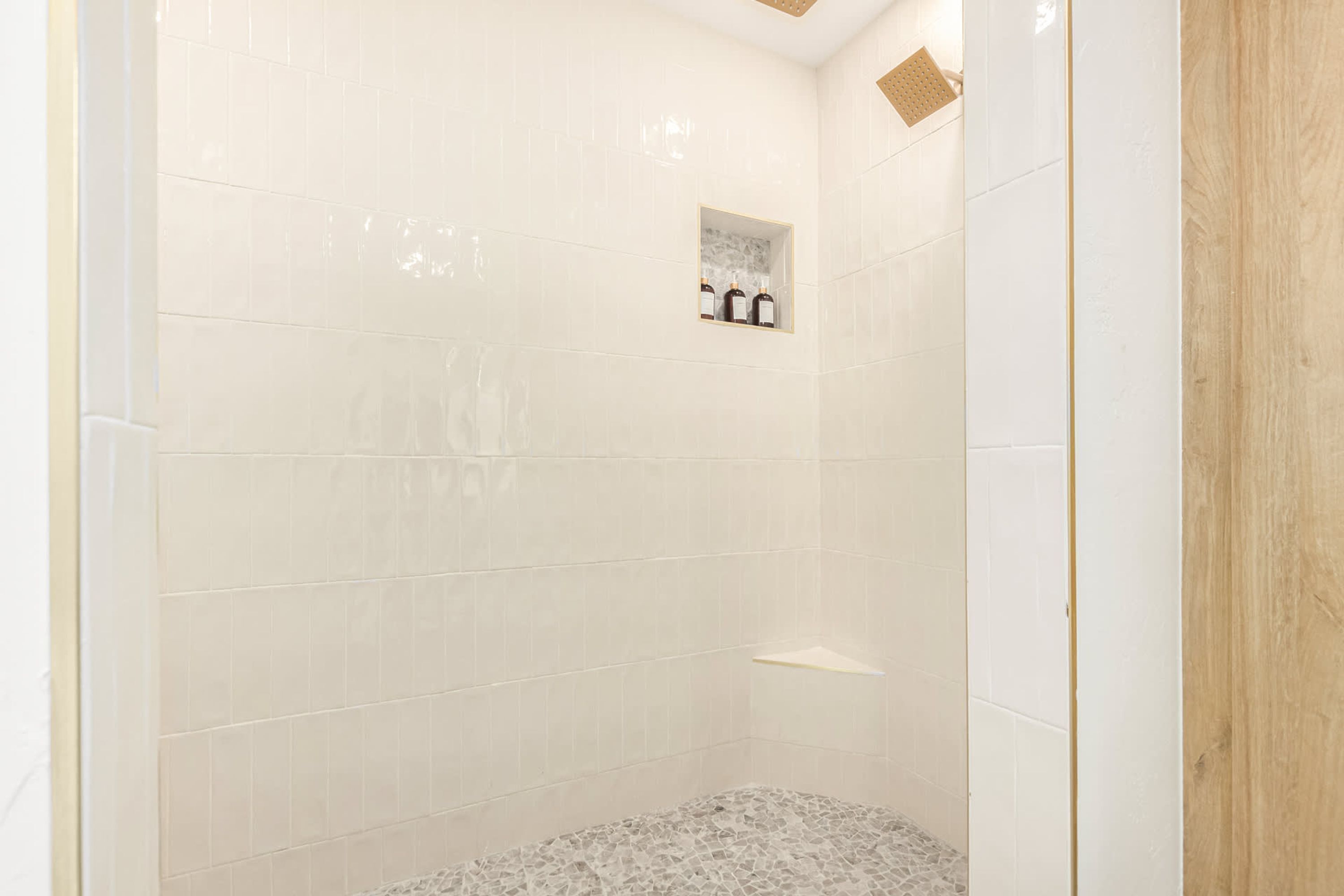 A shower area with white tiled walls, a small shelf with bottles, and a textured floor.