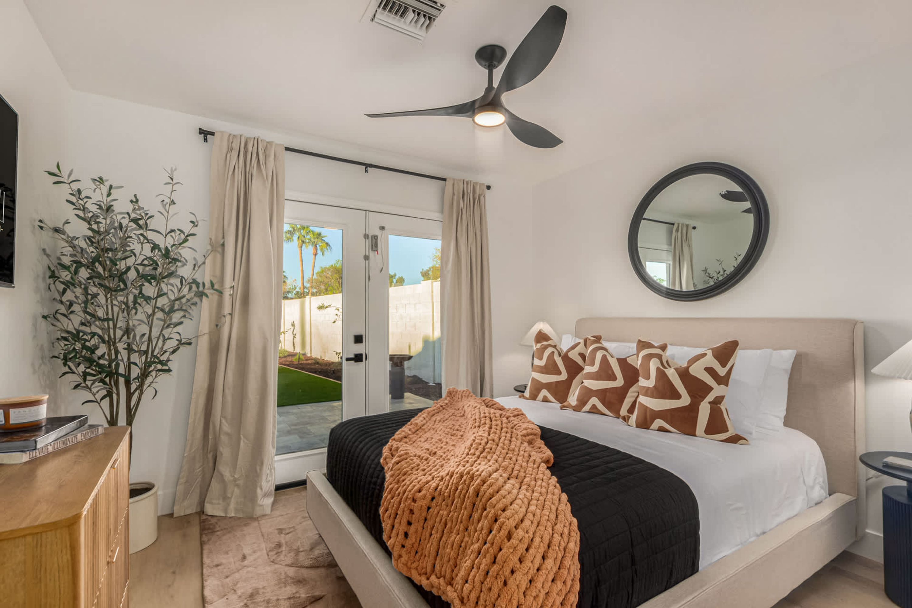 A bedroom with a bed, a ceiling fan, a round mirror, and a view through a glass door.