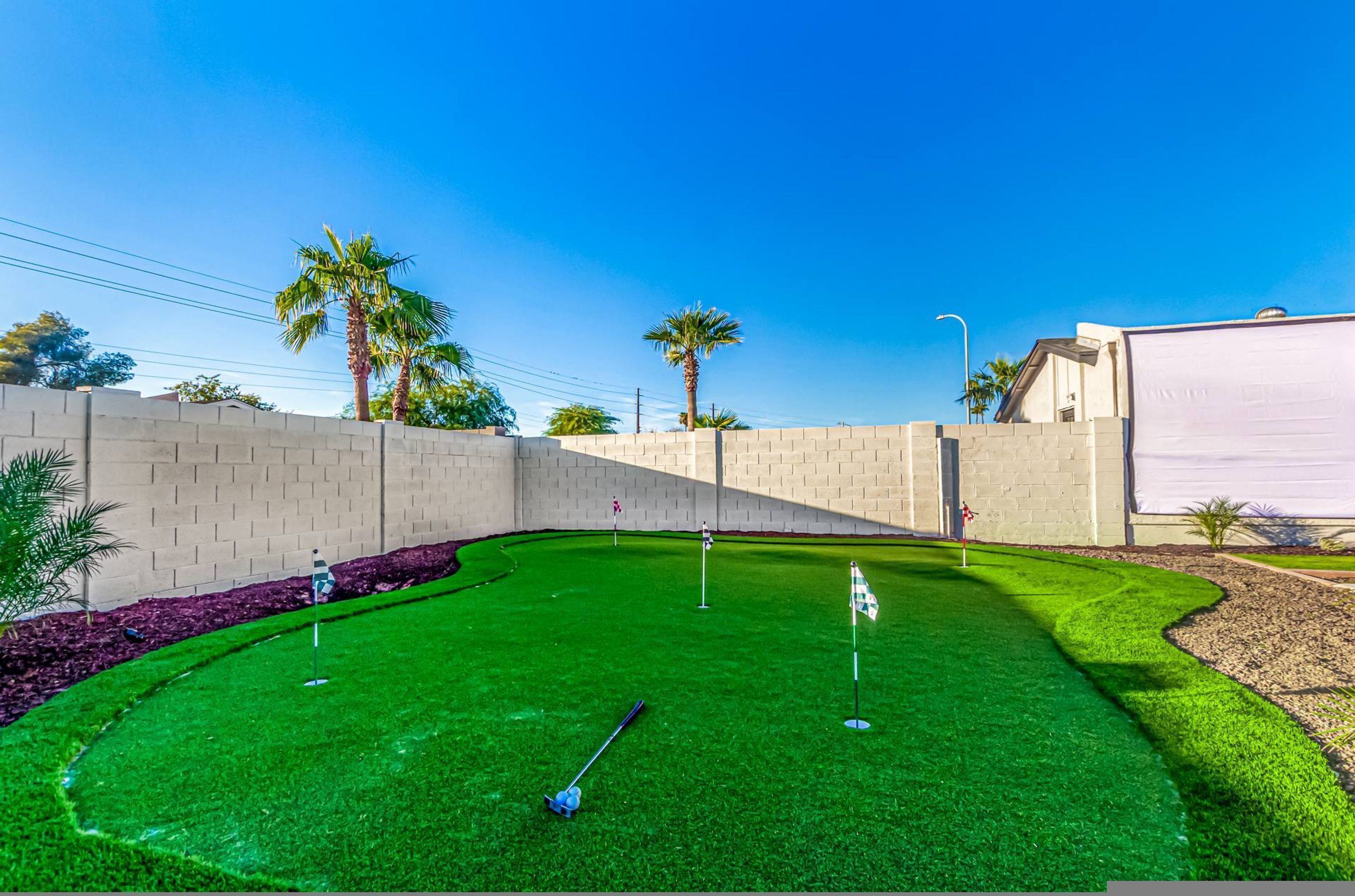 A well-maintained artificial green golf course with a brick wall, palm trees, and a clear blue sky.