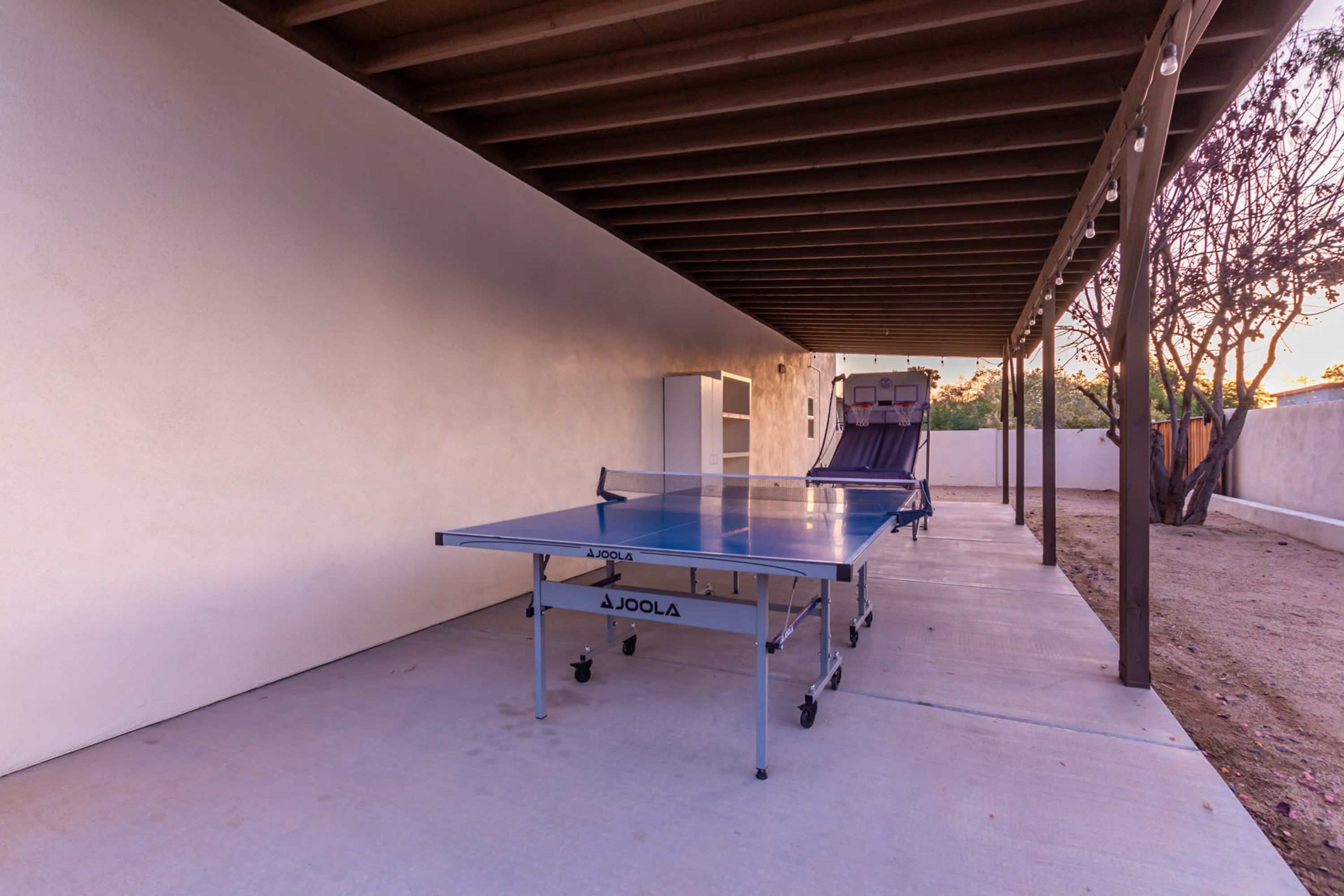 Una mesa de ping pong al aire libre bajo una pérgola de madera, con piso de concreto, rodeada por un muro bajo y árboles.