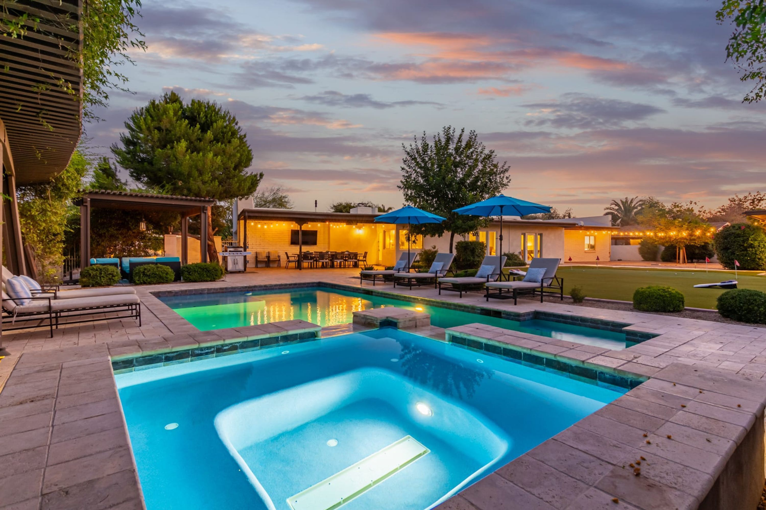 A swimming pool with lounge chairs and umbrellas, surrounded by a patio area with a covered seating area and string lights.