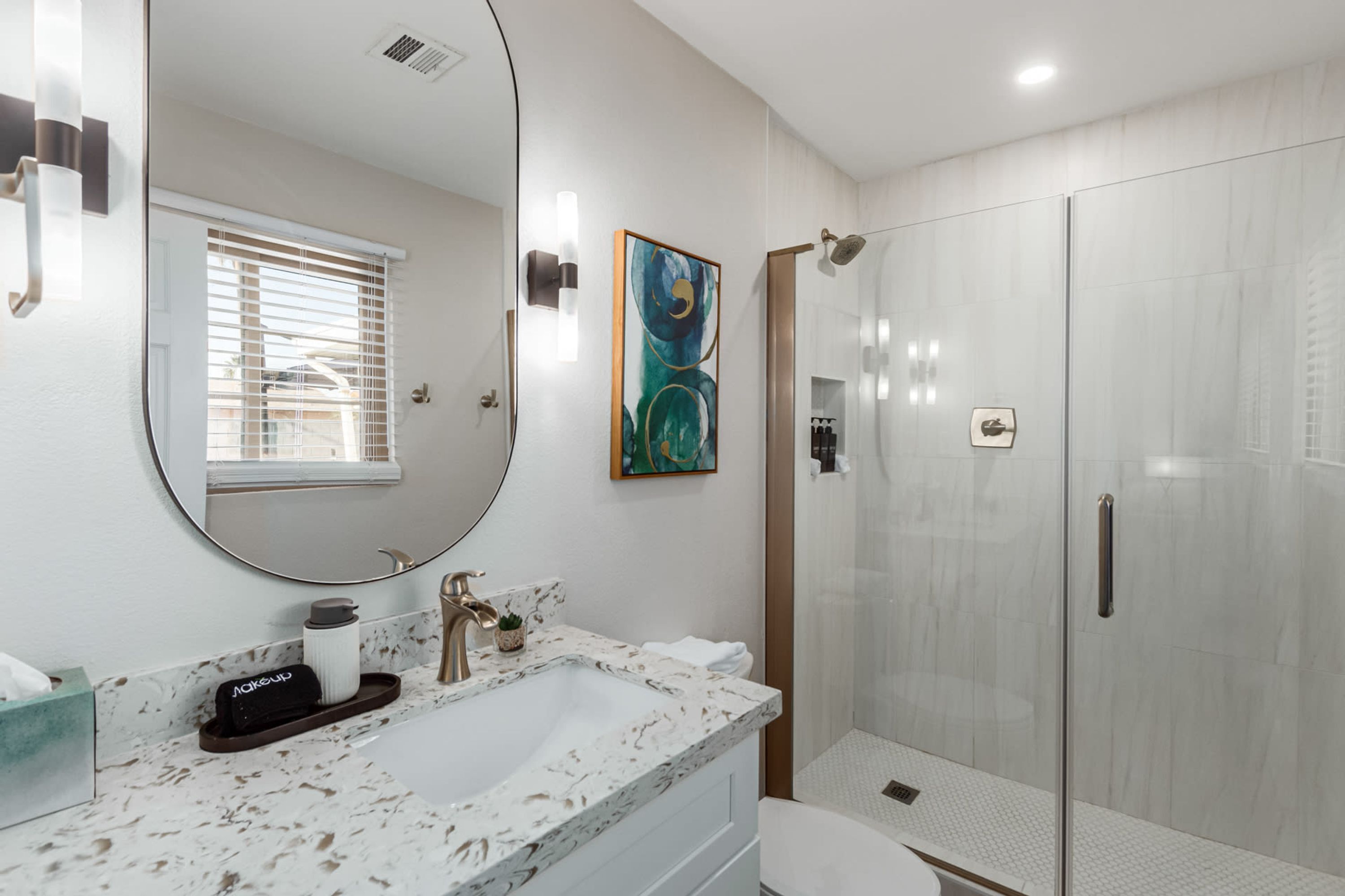 Un baño moderno con lavamanos de mármol, un espejo grande, cabina de ducha de vidrio y una obra de arte enmarcada en la pared.