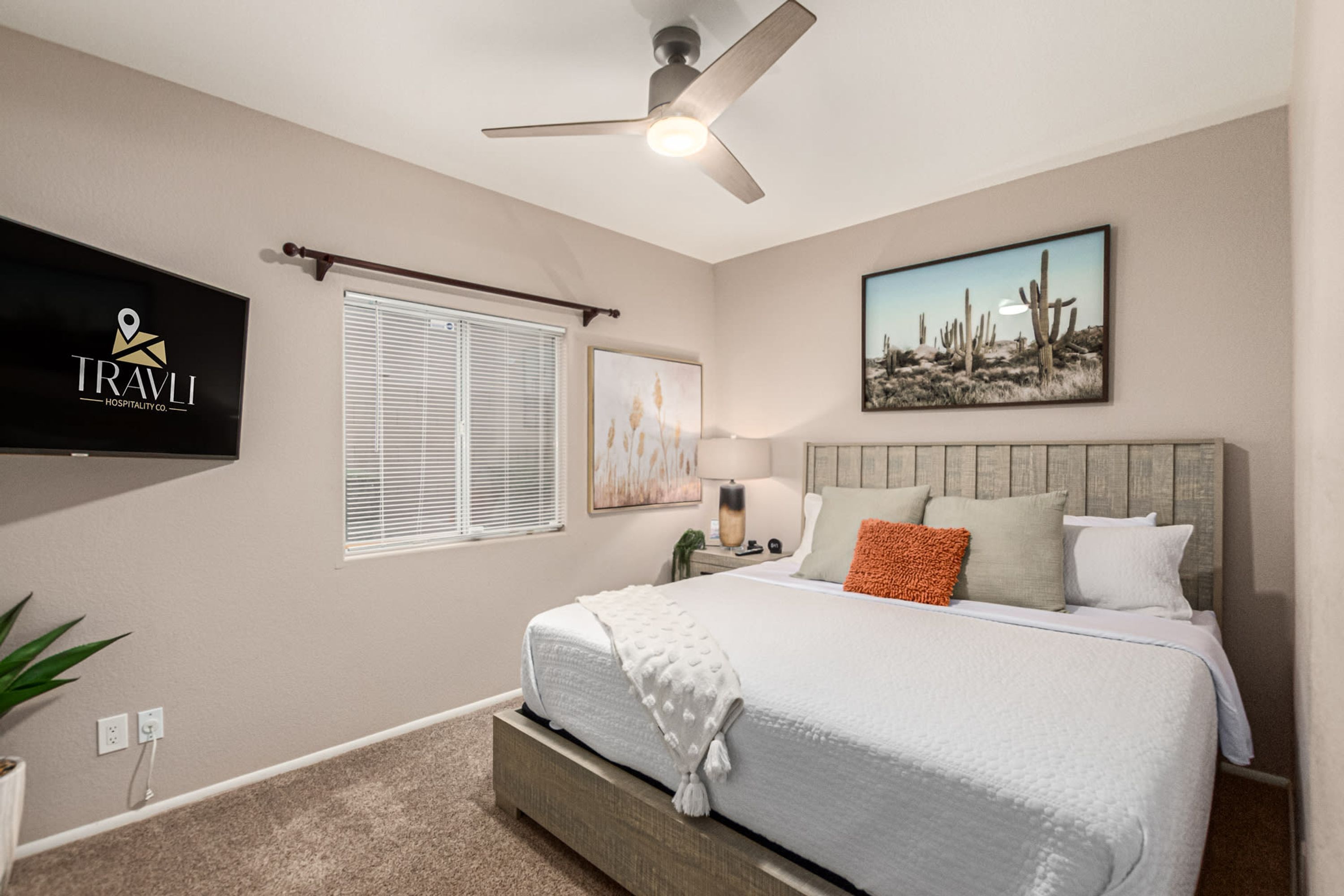 A hotel room with a bed, a ceiling fan, a TV, a window with blinds, and a desert-themed painting.