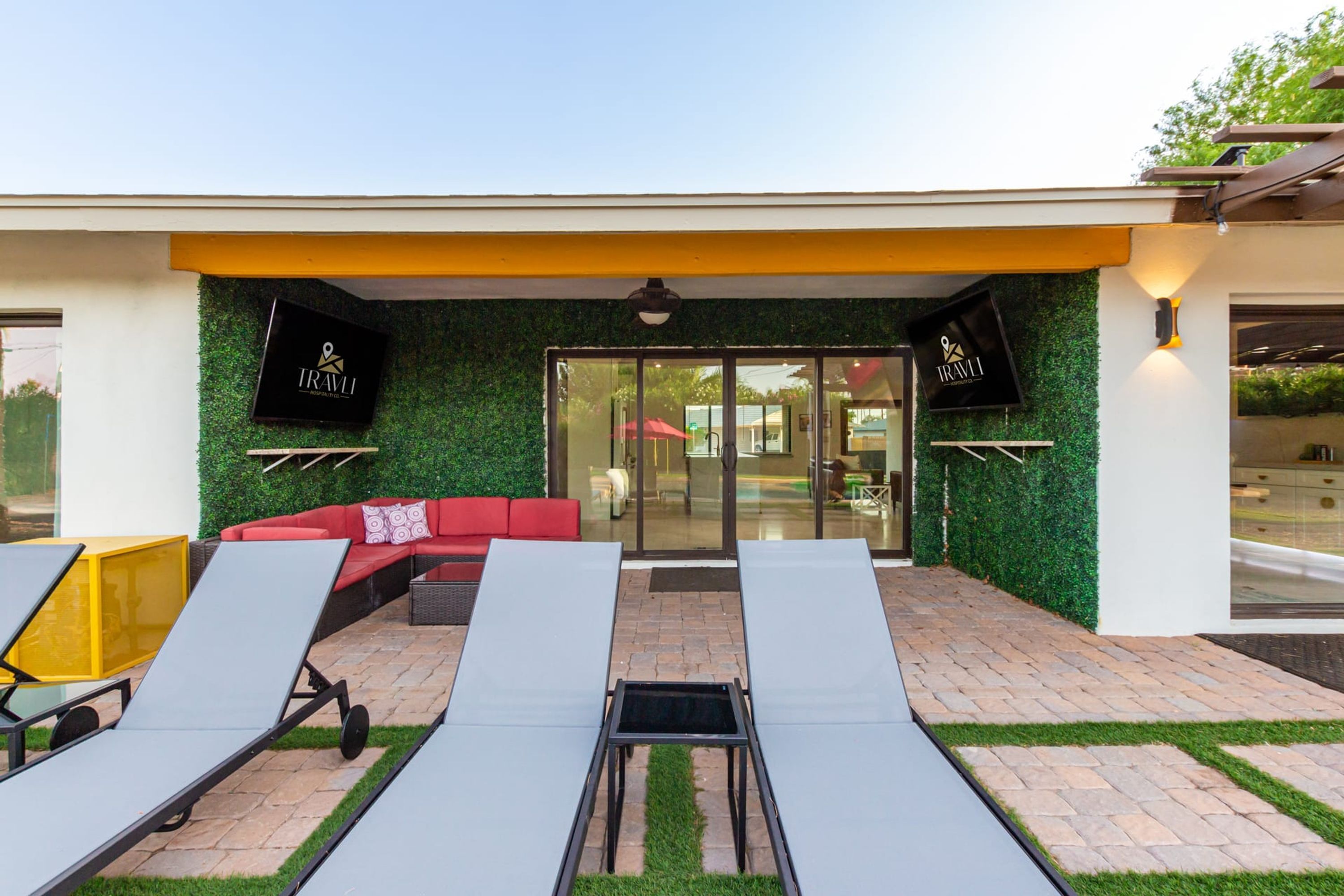 A modern outdoor lounge area with sun loungers, a red sofa, and a glass door entrance.