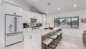 A modern kitchen with white cabinetry, a marble island, and a large window with blinds.