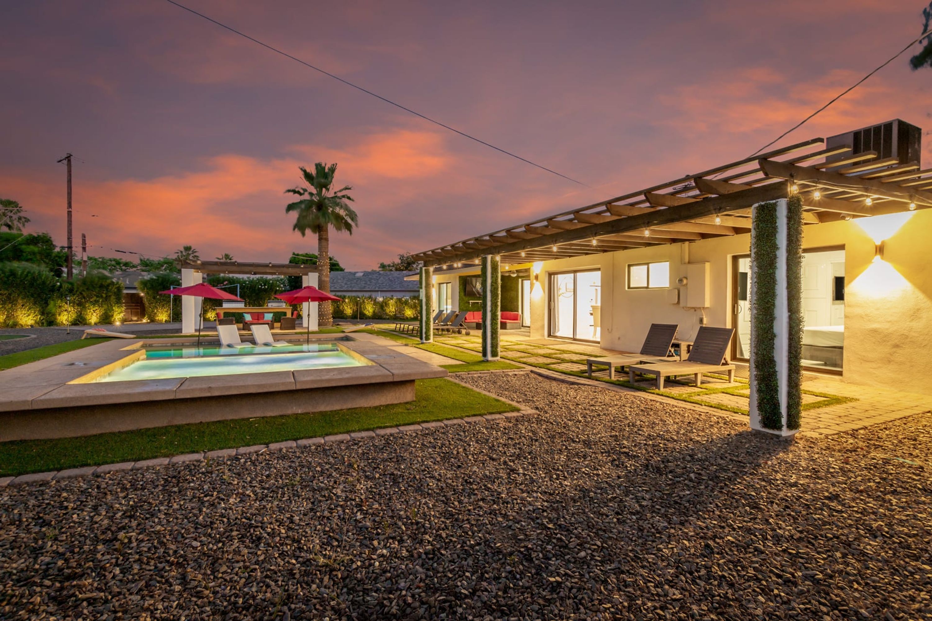 A modern outdoor pool area with lounge chairs and umbrellas, surrounded by a well-maintained garden and a building with large windows.