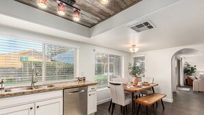 A modern kitchen with white cabinetry, a granite countertop, and a built-in dishwasher. There is a dining area with a wooden table and chairs, and a large window with blinds. - Spanish Wells Retreat Free Pool Heat Spa BBQ (Scottsdale)