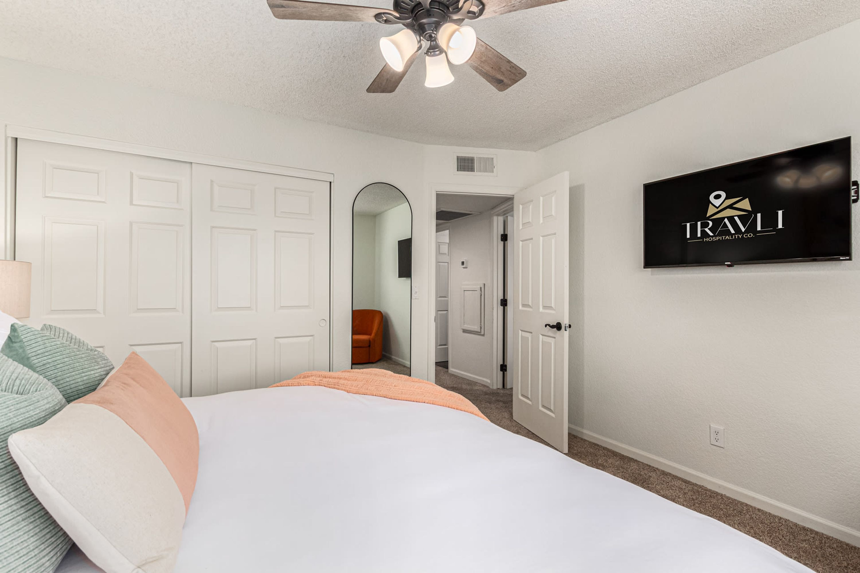 A bedroom with a bed, white closet doors, a ceiling fan, and a TV displaying 'TRAVLI HOSPITALITY CO.'