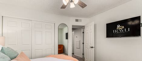 A bedroom with a bed, white closet doors, a ceiling fan, and a TV displaying 'TRAVLI HOSPITALITY CO.'