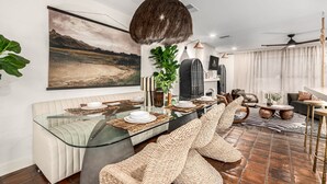A modern dining area with a glass table, cushioned seating, and a large landscape painting on the wall.
