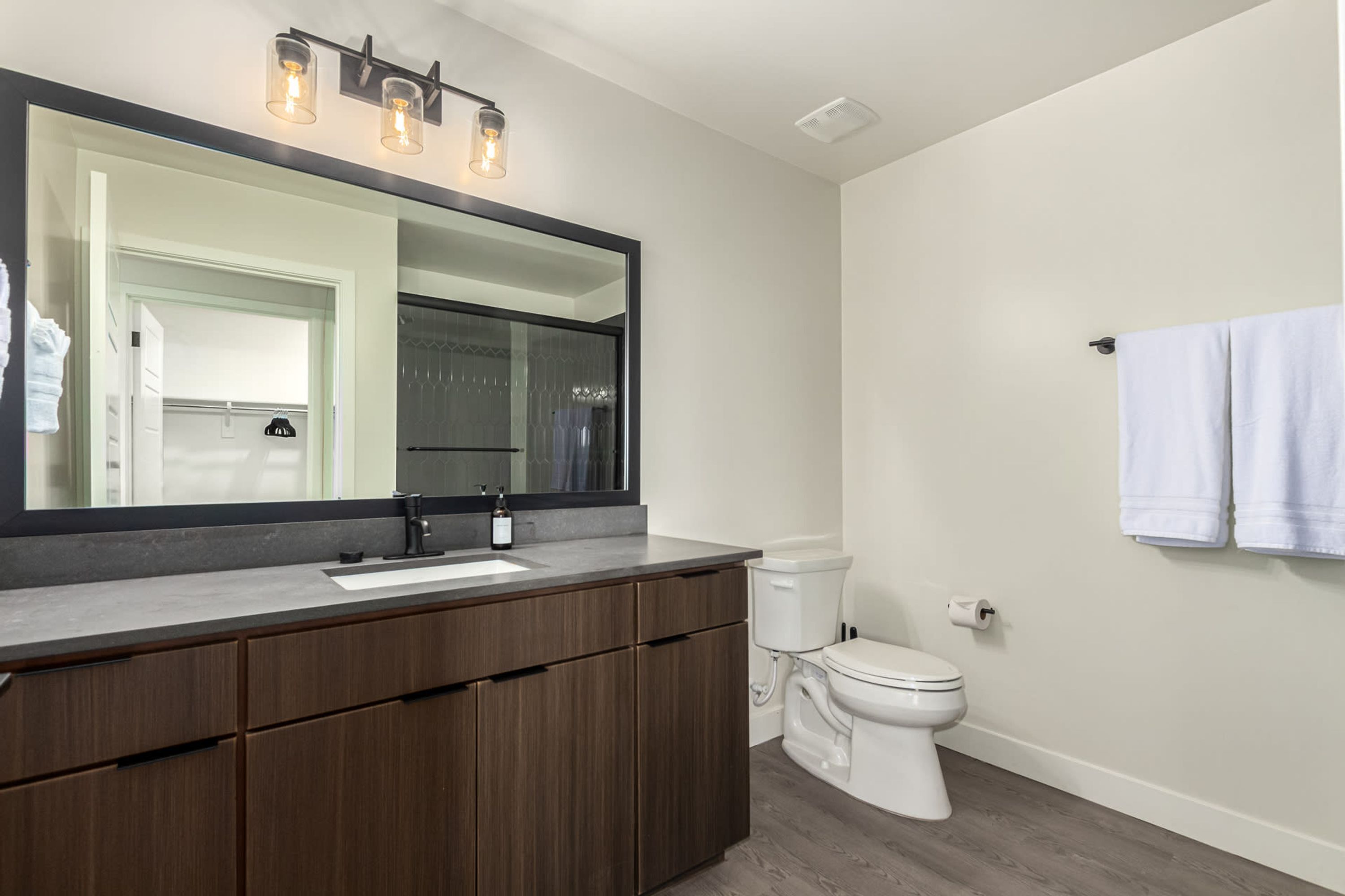 Une salle de bain moderne avec un grand miroir, un meuble-lavabo en bois foncé, des toilettes blanches et un porte-serviettes.