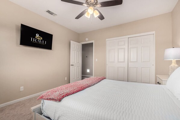 A bedroom with a bed, a ceiling fan, a TV mounted on the wall, and a door leading to another room.