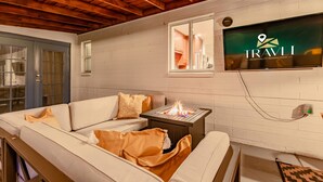 A cozy room with a white brick wall, a flat-screen TV mounted above a sofa, and a small table with a fire feature. - Desert Diamond Oasis Private Pool Bar Games (Scottsdale)